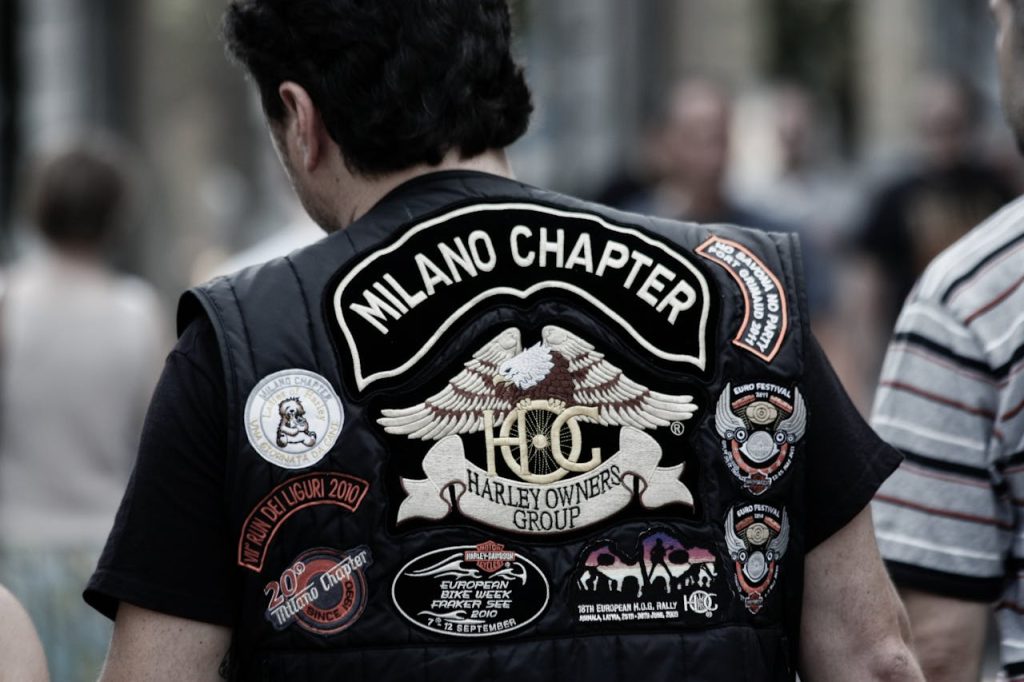 Home Close-up of a biker wearing a Milano Chapter Harley Owners Group vest adorned with patches.